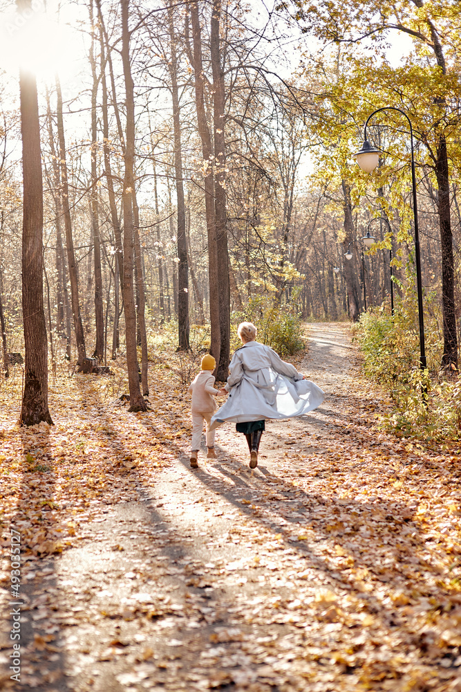 Fototapeta premium Rear view on cheerful mother and child boy having fun in autumn park, running, having rest. friendly family in casual clothes walking, spending time together, at summer day. view from back