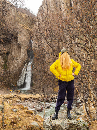 A woman in a yellow jacket with hair instead of a face stands against the backdrop of rocks and a waterfall in an autumn mountain gorge. Comic image