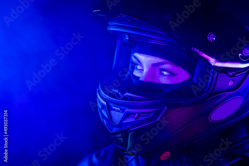 Portrait of confident motorcyclist woman in closed motorcycle helmet. Young driver biker under blue neon light at night. Copy space.