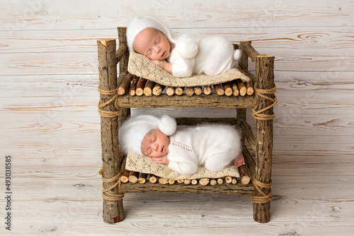 Tiny newborn twin boys in white bodysuits against a light wood background. Newborn twins sleep on a bunk wooden bed. The boys are dressed in white caps. Professional studio photography.