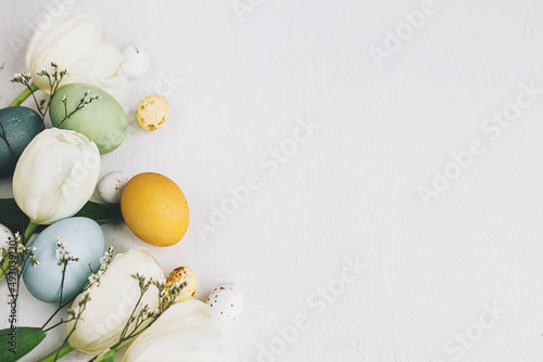 Stylish Easter eggs and tulips border on rustic white wooden background, flat lay with space for text. Happy Easter! Natural dyed colorful eggs and spring flowers composition. Greeting card photo