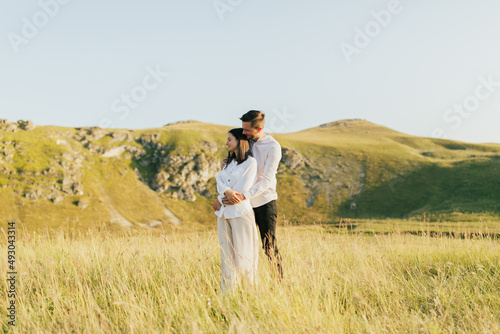 Man hugging his woman from behind while standing on the meadow with mountains on the background. Romantic moment, summer relaxing together concept. © eduard