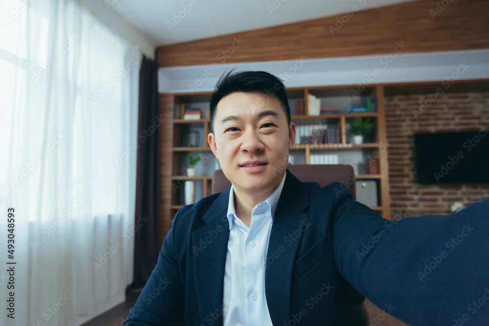 Cheerful and funny Asian businessman looks at the camera of a smartphone, talks on a video call, a man works in the office, waves a congratulatory gesture Cheerful and funny Asian businessman looks at the camera of a smartphone, talks on a video call, a man works in the office, waves a congratulatory gesture