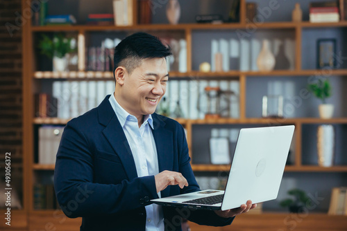 Happy asian freelance businessman working standing, holding laptop, smiling and rejoicing, at home office