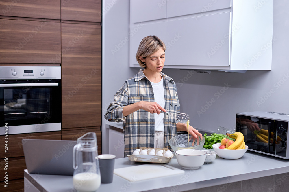Beautiful, attractive young woman kneading dough with whisk for banana pancakes in bowl in kitchen with white, light interior. Caucasian female in casual checkered shirt at home alone