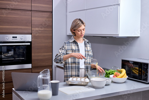 Beautiful  attractive young woman kneading dough with whisk for banana pancakes in bowl in kitchen with white  light interior. Caucasian female in casual checkered shirt at home alone