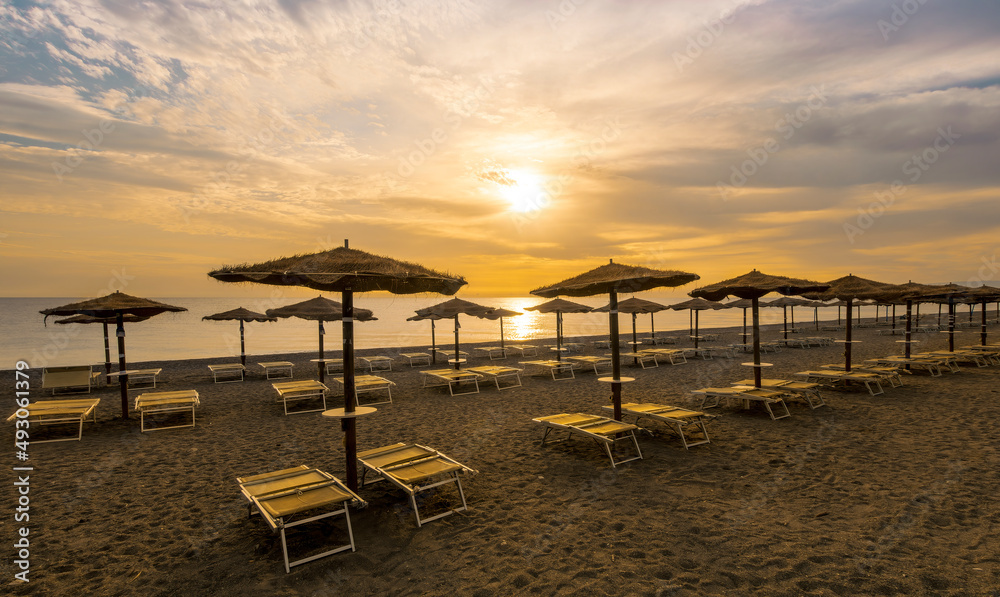  empty beach during beautiful sunrise or sunset with chaise loungues and nice umbrellas with blue sea, sun glow and amazing cloudy sky on thr background
