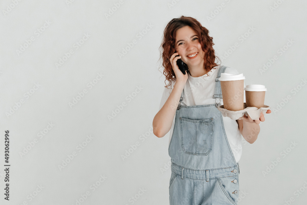 Excited redhead girl, wearing denim dungarees and white t-shirt, holding takeaway coffee cups, talking to her friend on the phone, over white background Excited redhead girl, wearing denim dungarees and white t-shirt, holding takeaway coffee cups, talking to her friend on the phone, over white background
