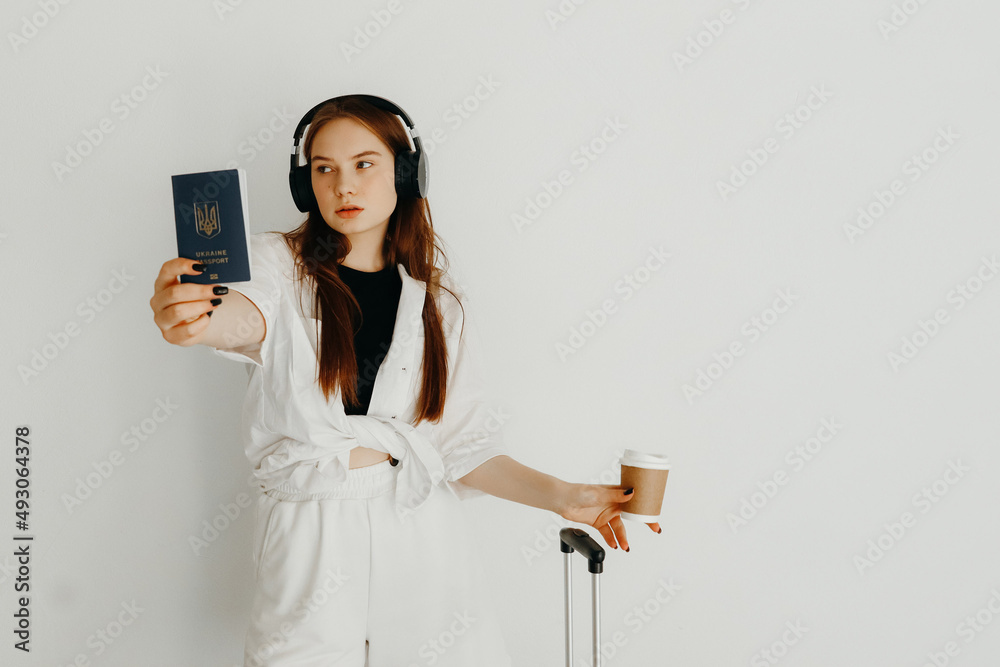 Naklejka premium Young woman with cup of coffee, headphones, and a suitcase showing her passport, on a white background. Travel concept. Woman is going to travel by plane