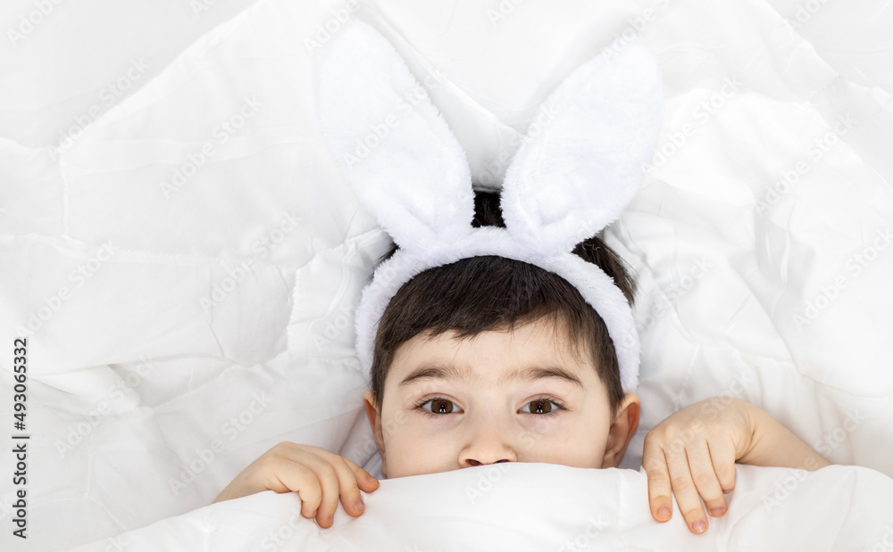 Happy Easter concept. cute beautiful kid, boy, with green and brown color eyes and white bunny ears, is peeking out from under the white blanket. minimalist photo Happy Easter concept. cute beautiful kid, boy, with green and brown color eyes and white bunny ears, is peeking out from under the white blanket. minimalist photo