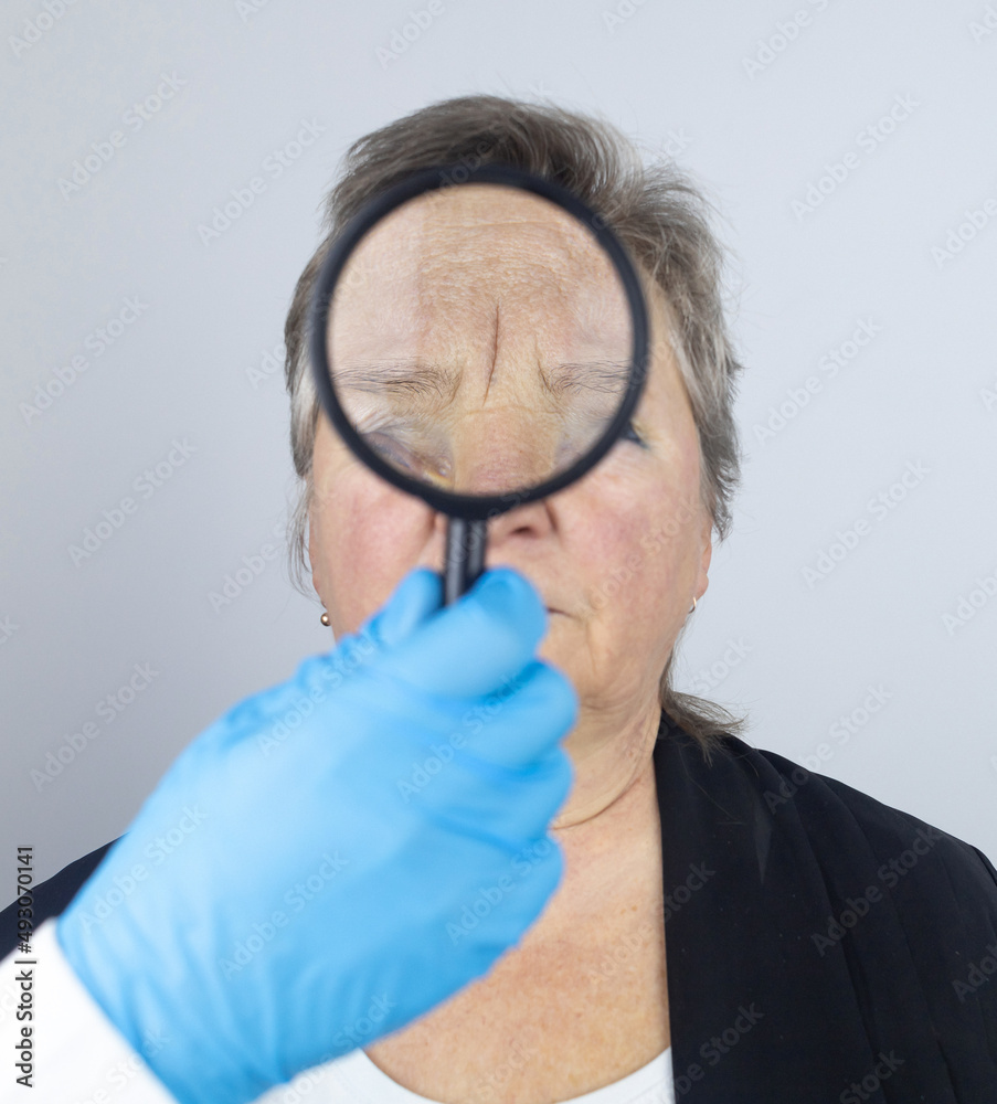 Senior woman at the reception of a facial plastic surgeon. Consultation on the removal of age wrinkles on the face. Doctor shows what procedures he will do. Mimic wrinkles. Magnifying glass in hands Senior woman at the reception of a facial plastic surgeon. Consultation on the removal of age wrinkles on the face. Doctor shows what procedures he will do. Mimic wrinkles. Magnifying glass in hands