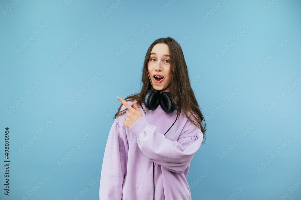 A funny lady with a happy happy face points her finger to the copy space and looks at the camera, wearing a sweater on headphones. Isolated on a blue background A funny lady with a happy happy face points her finger to the copy space and looks at the camera, wearing a sweater on headphones. Isolated on a blue background