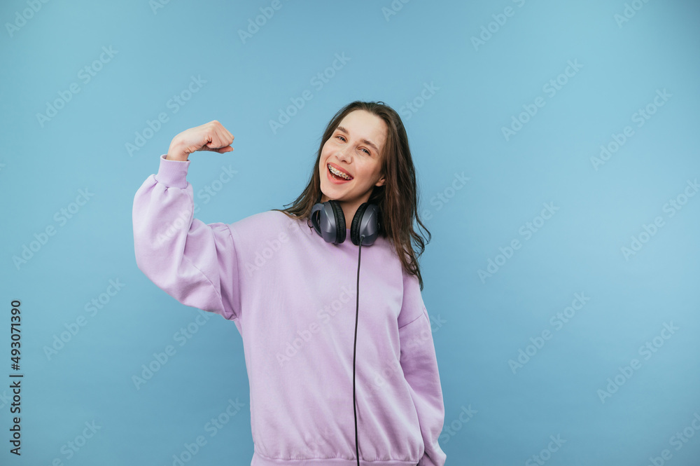 Attractive brunette girl in purple sweater and headphones shows biceps to camera and smiles. Attractive brunette girl in purple sweater and headphones shows biceps to camera and smiles.