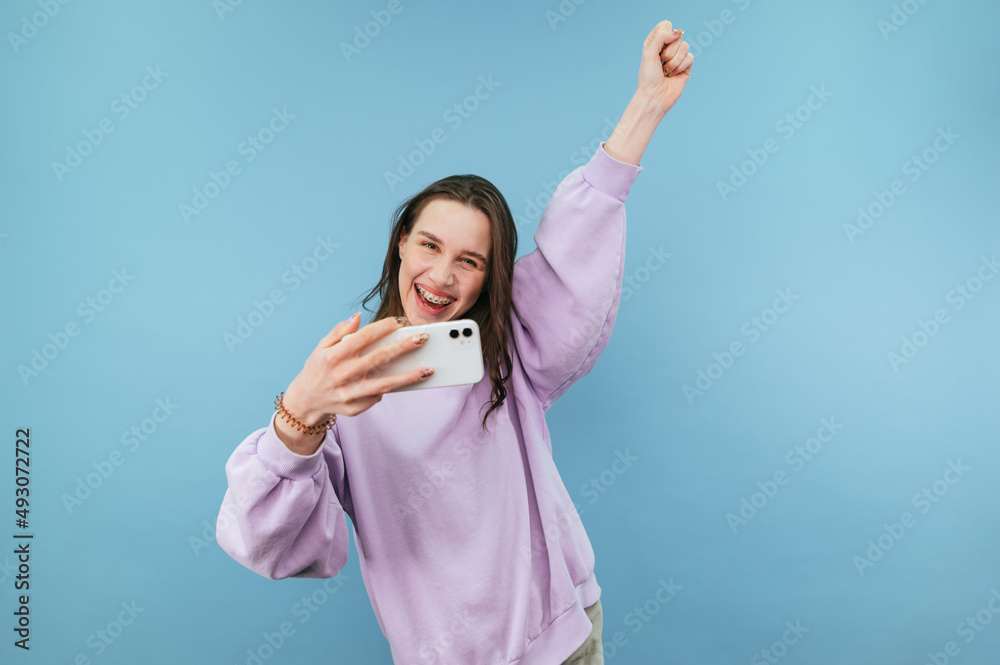 Joyful girl gamer with a smartphone in hand emotionally rejoices in winning an online game on a blue background, looks at the camera and smiles. Joyful girl gamer with a smartphone in hand emotionally rejoices in winning an online game on a blue background, looks at the camera and smiles.
