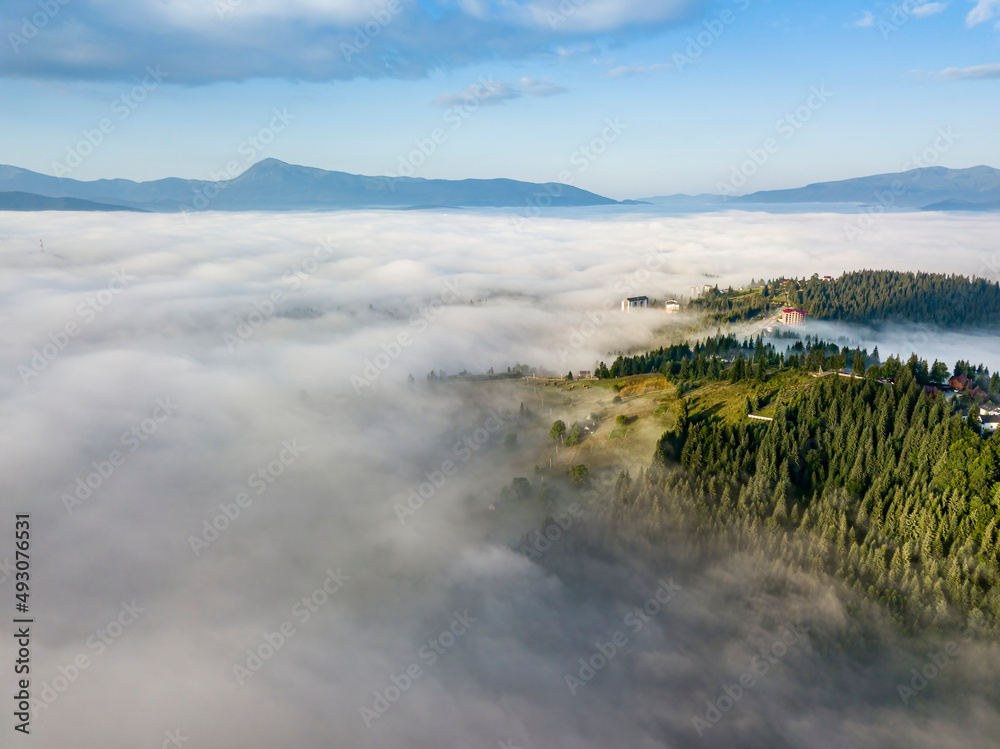 Fog envelops the mountain forest. The rays of the rising sun break through the fog. Aerial drone view.