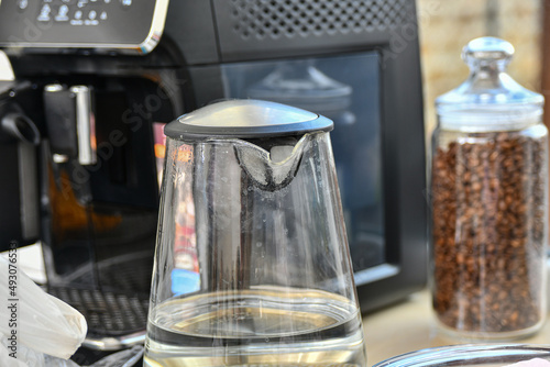 A glass kettle with hot water for coffee next to the coffee machine in the kitchen.