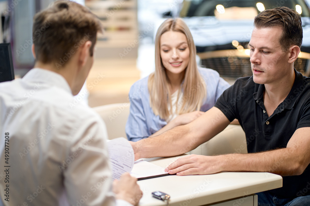 young male shop assistant explaining talking offering buying new car to young caucasian family husband and wife at auto dealer shop, sit talking discussing. in cars showroom young male shop assistant explaining talking offering buying new car to young caucasian family husband and wife at auto dealer shop, sit talking discussing. in cars showroom