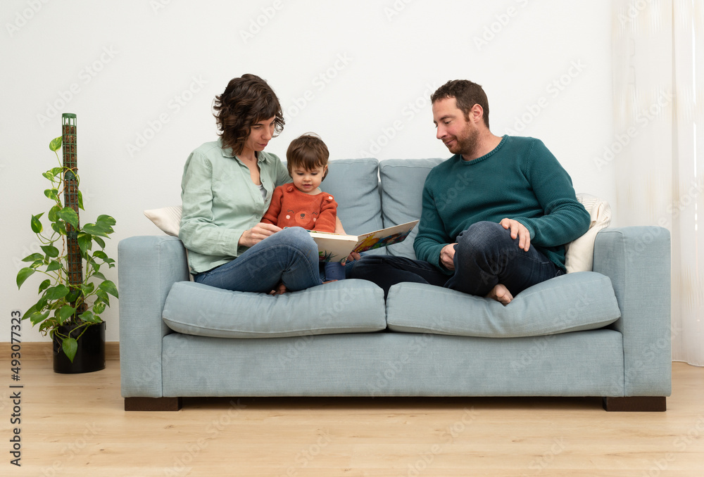 Family (mother, father and toddler) reading a book on a sofá. Time together. 