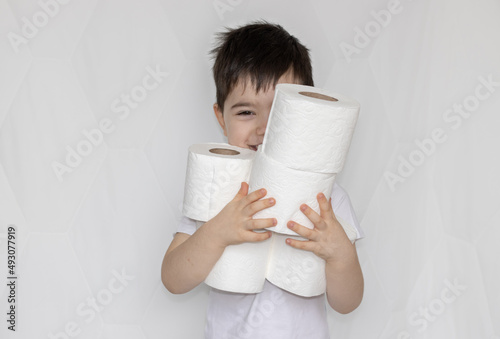 cute little boy is holding toilet paper rolls in arms, or is looking through the hole. stack of wc paper rolls. happy boy is having fun. child is smiling. hygiene. boy in white t shirt on grey back
