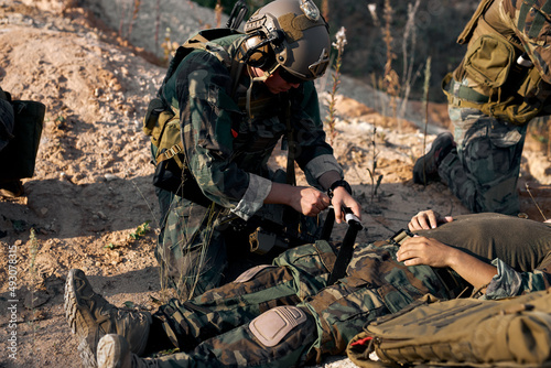 Foto young caucasian military man bandages a leg of injured comrade on fight battlefield, lying on ground