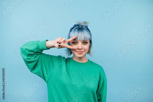 Attractive lady with blue hair poses for the camera with a smile on her face and shows a gesture of peace on a blue background.