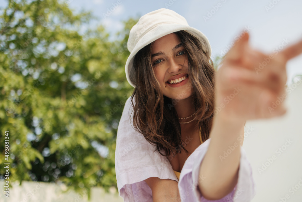Close-up of cute young caucasian brunette woman smiling with teeth at camera on blurry street background. Girl in panama hat dressed light summer clothes. Vacation, happiness and pleasure concept