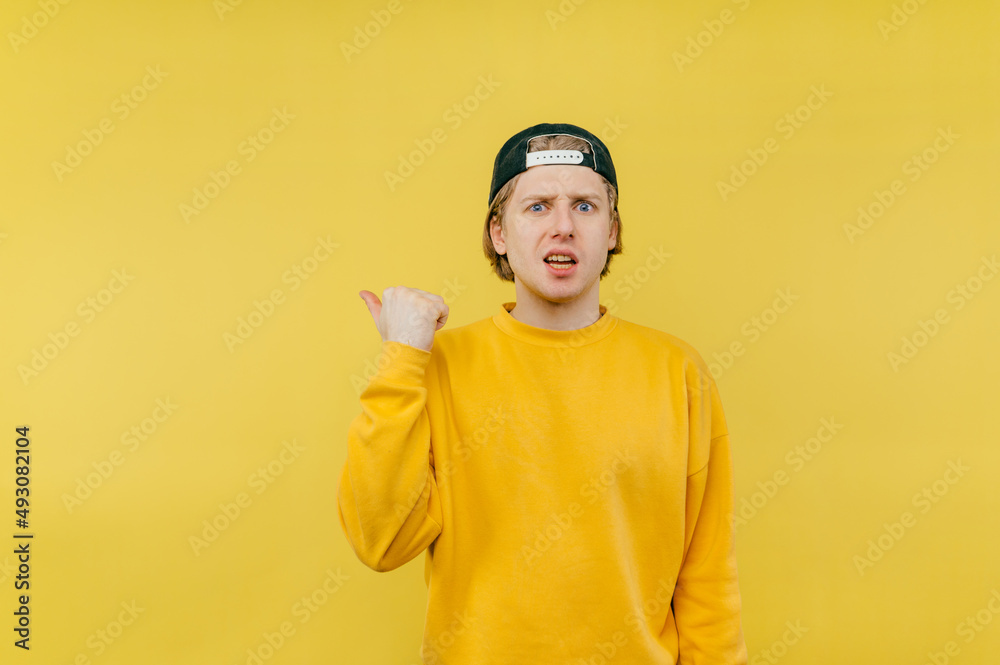 Emotional disgruntled guy in a cap and sweatshirt points his finger to the copy space and looks at the camera on a yellow background. Emotional disgruntled guy in a cap and sweatshirt points his finger to the copy space and looks at the camera on a yellow background.
