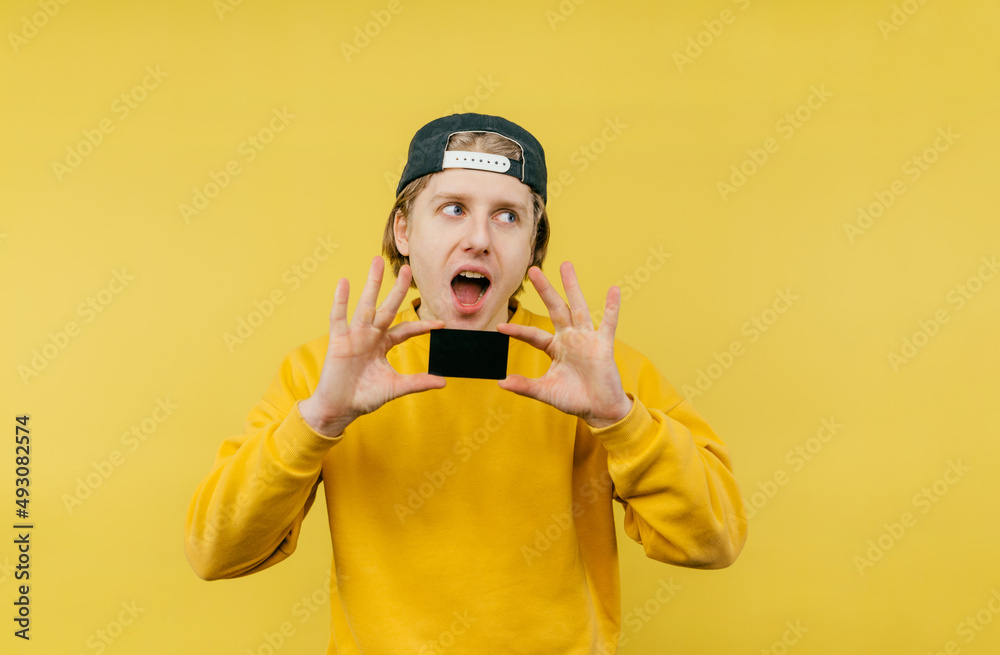 Joyful young man with a bank card in his hands stands on a yellow background and looks away with a surprised face.