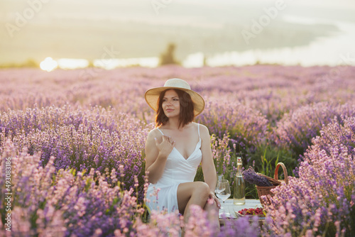 Fototapeta Naklejka Na Ścianę i Meble -  Elegant woman in white dress and straw hat on the lavender field having picnic at sunset. A girl on a picnic in the flowering lavender. River on the background.