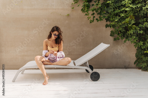 Portrait of cute young caucasian woman sitting on sun lounger in sunglasses against grey wall. Brunette with loose hair is resting on weekend. Summer lifestyle concept.