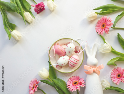 Layout of easter table decoration with easter bunny and pink eggs on roses plate, with pink daisy flowers and white tulips. Easter bunny with ribbon. Celebration layout or card. Minimalist table decor