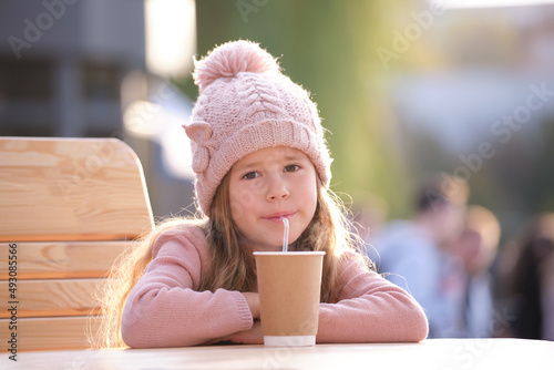 Portrait of cute little child girl in pink hat sitting alone at street cafe drinking tea from paper cup