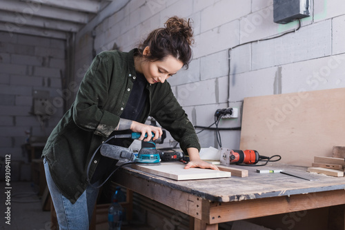 Carpenter in shirt sanding wooden plank in workshop.