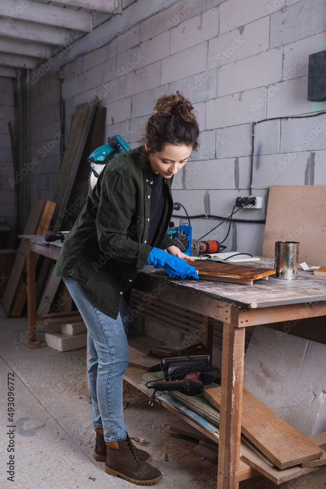 Young carpenter in rubber glove applying wood stain on plank near jar and miter saw in workshop. Young carpenter in rubber glove applying wood stain on plank near jar and miter saw in workshop.