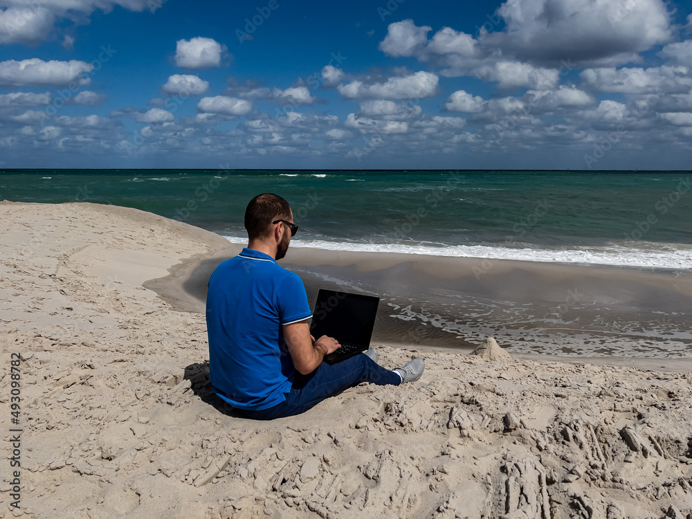 A brunette man in a blue t-shirt works with a computer on the ocean. Horizontal photo. Soft focus. 