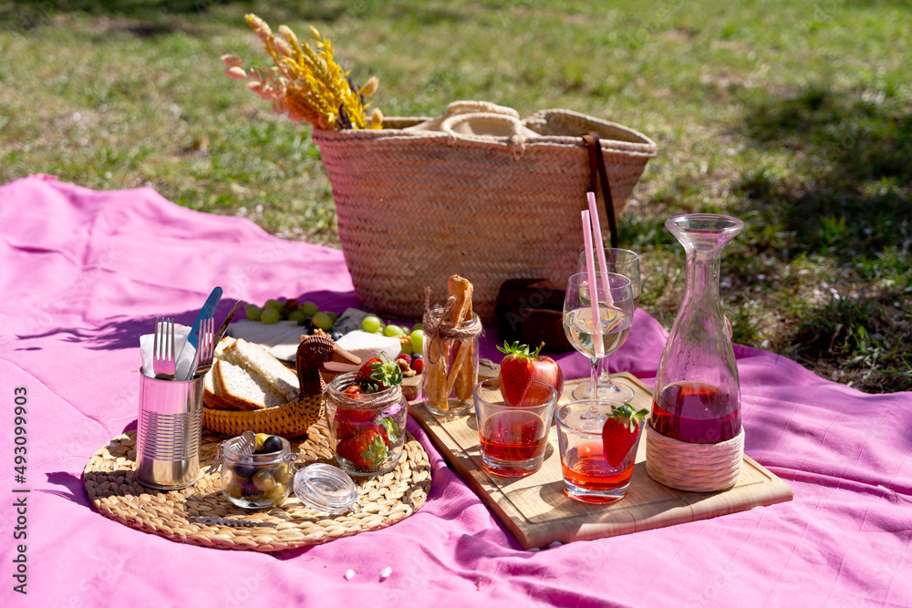 Panoramic side view of bohemian picnic outdoor with wicker basket in the park. Horizontal full length image of variety of snack food set with pink blanket background. Gastronomy and food concept.