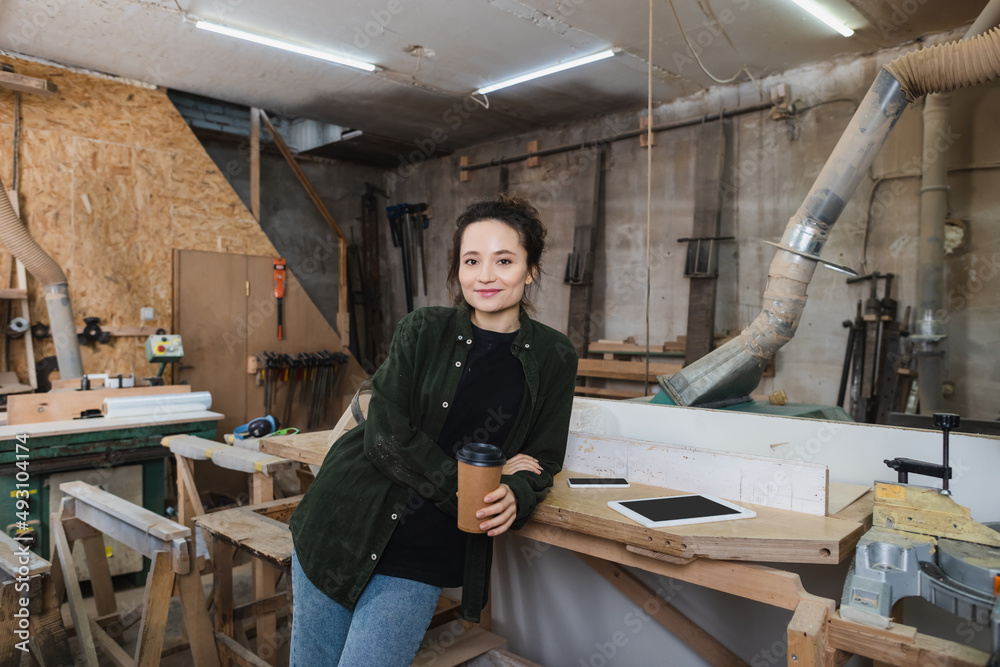 Cheerful carpenter holding coffee to go and looking at camera in workshop. Cheerful carpenter holding coffee to go and looking at camera in workshop.