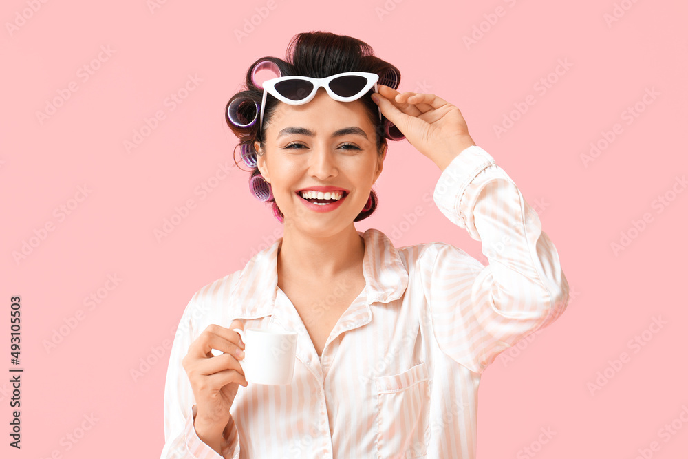Beautiful young woman in pajamas, hair curlers and with sunglasses drinking coffee on pink background Beautiful young woman in pajamas, hair curlers and with sunglasses drinking coffee on pink background