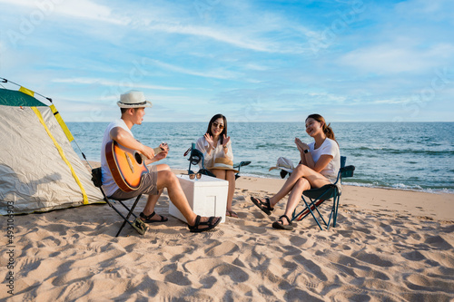 Group of happy Asian friends playing guitar and singing with clap while picnic and camping on the beach in outdoors vacation summer.
