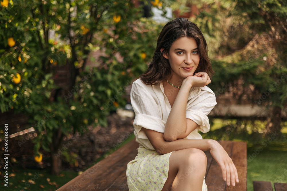 Young pretty fair-skinned lady is playfully looking at camera while sitting in orchard alone. Dark-haired model wears light-colored casual summer clothes. Lifestyle, emotions, leisure concept. Young pretty fair-skinned lady is playfully looking at camera while sitting in orchard alone. Dark-haired model wears light-colored casual summer clothes. Lifestyle, emotions, leisure concept.