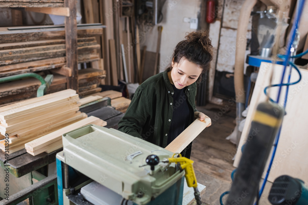 Brunette carpenter holding wooden board near thickness planer and planks in workshop.