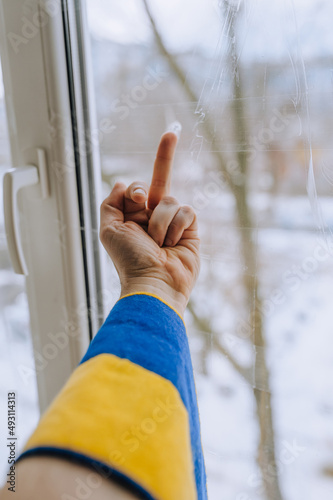 A Ukrainian man, being at home near a window in Ukraine under shelling in the war, shows an obscene gesture, the middle finger to the Russian occupiers. Concept, conflict, photography.