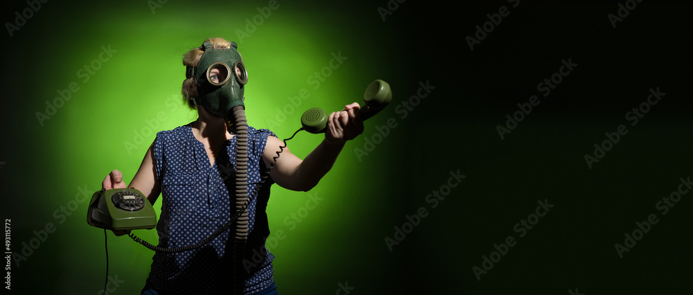 A woman in a gas mask passes the receiver of a retro phone on a dark dramatic background with copy space, hard light.