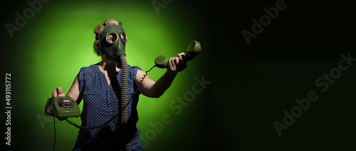 A woman in a gas mask passes the receiver of a retro phone on a dark dramatic background with copy space, hard light. © Sergey
