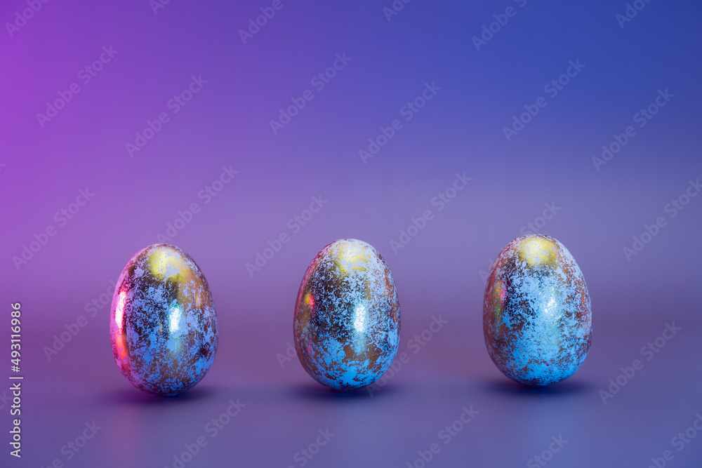 Row of three Easter golden eggs with different patterns in neon colors light. Purple very peri color minimalist festive background. Modern trendy Easter. Selective focus. Copy space. Row of three Easter golden eggs with different patterns in neon colors light. Purple very peri color minimalist festive background. Modern trendy Easter. Selective focus. Copy space.
