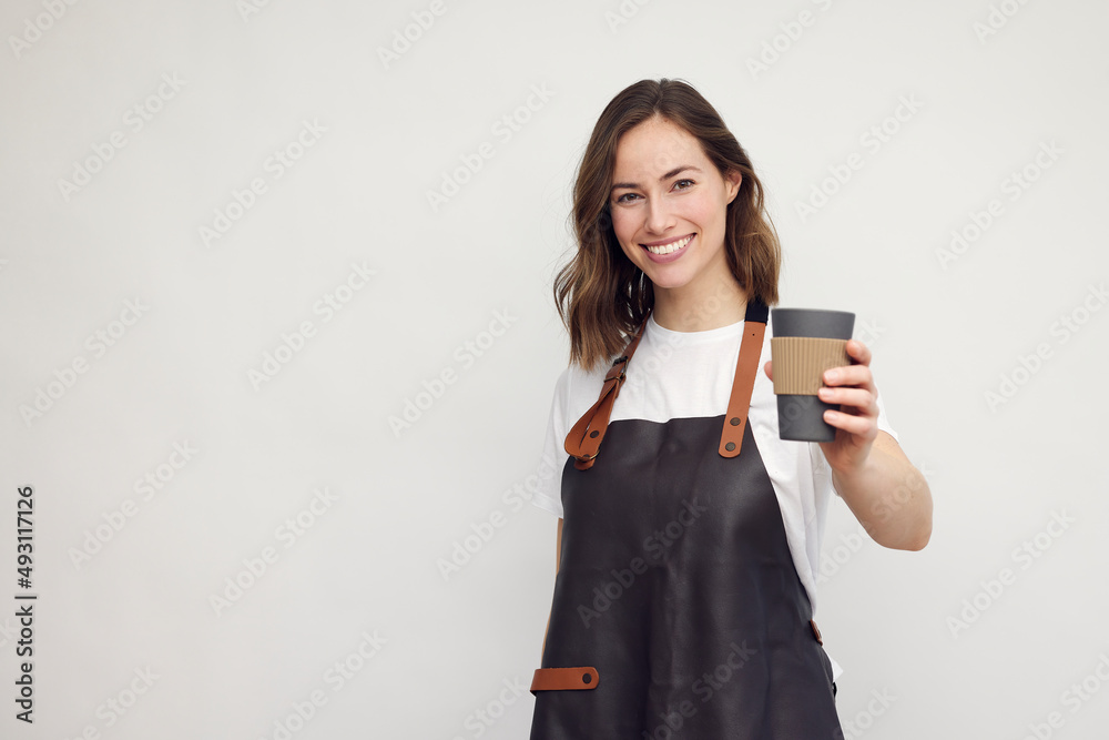 Fototapeta premium Smiling young barista woman in apron isolated on white background. Looking beautiful and happy with a to-go coffee cup in hand. Concept: serving coffee.