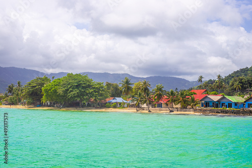 Tropical seascape. Palm trees leaned over the water, next to the roofs of houses. View from the sea to the coastal strip