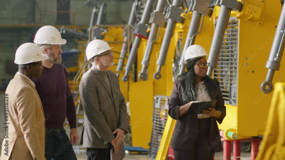 Young African American female manager in formalwear and hardhat telling about heavy equipment and giving clipboard to group of senior multiethnic businessmen during their visit to factory