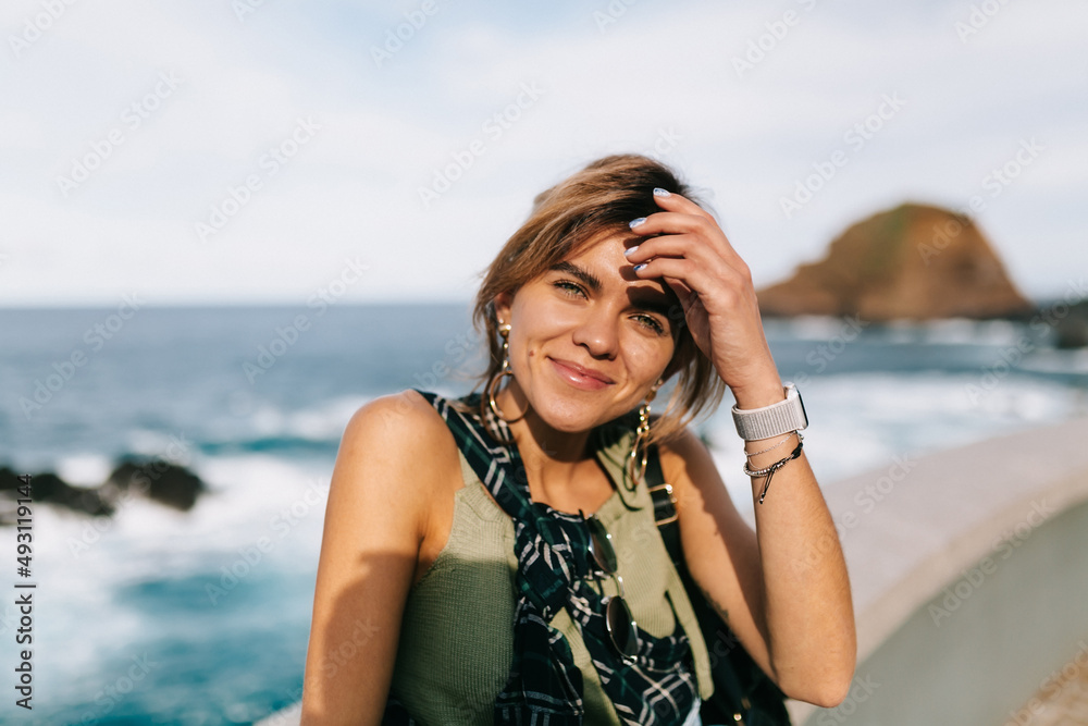 Close up outside portrait of young woman is posing at camera while sitting on the ocean background in sunlight. Travel woman on the shore near the ocean Close up outside portrait of young woman is posing at camera while sitting on the ocean background in sunlight. Travel woman on the shore near the ocean