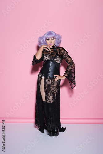 full length view of transgender person in black lace dress and violet wig on pink.